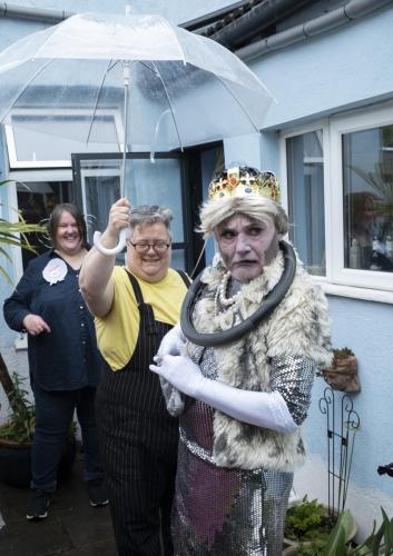 Aaron Williamson in drag as the ghost of queen diana, gaudy makeup, royal garb, a steering wheel round her neck, irritable expression. She is protected from the rain by a royal umbrella holder. A fat dyke laughs in the background.